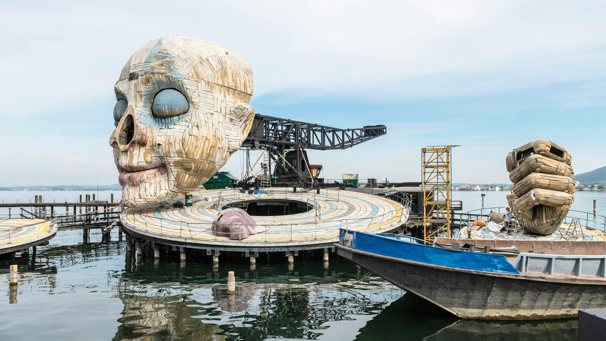 Large artistic installation resembling a human skull on a circular platform by the water, with boats nearby.