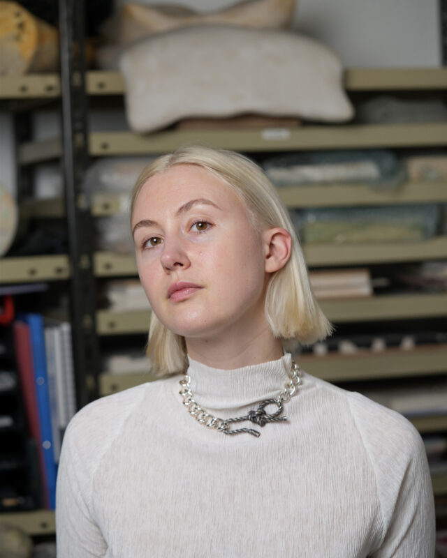 Portrait of Laura Dominici in a studio, wearing a light turtleneck and a statement necklace, with shelves of art materials...