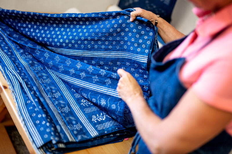 A person in a pink shirt holds a folded blue fabric with intricate white patterns, showcasing textile craftsmanship.