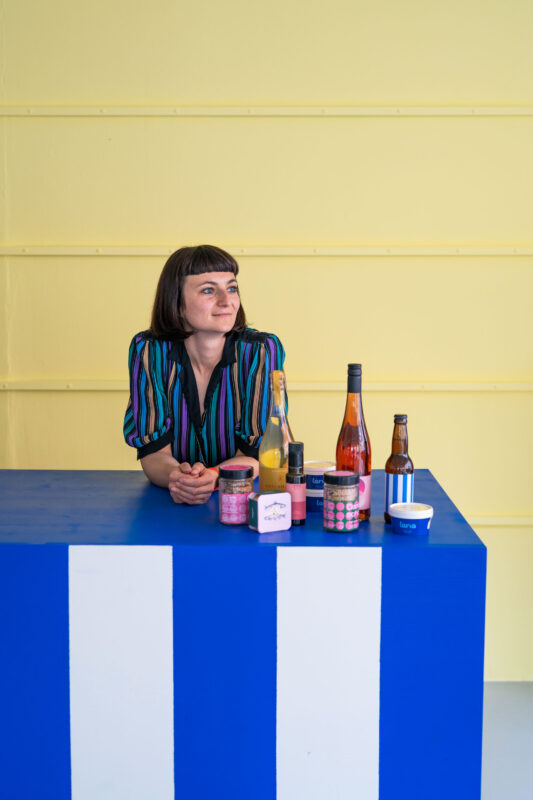 Woman in a striped shirt leans on a blue and white table displaying various beverages against a yellow background.
