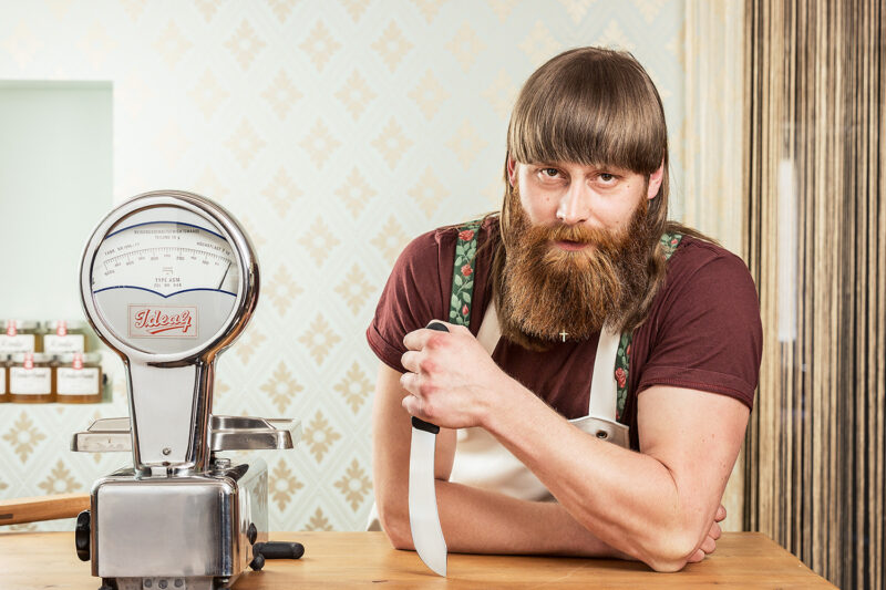Bearded man in a brown shirt and apron leans on a meat slicer, holding a knife, in a kitchen setting.