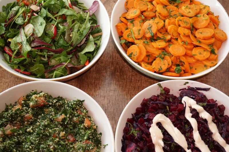 Four bowls of colorful salads: mixed greens, carrot salad, tabbouleh, and red cabbage with sauce, arranged on a wooden table.