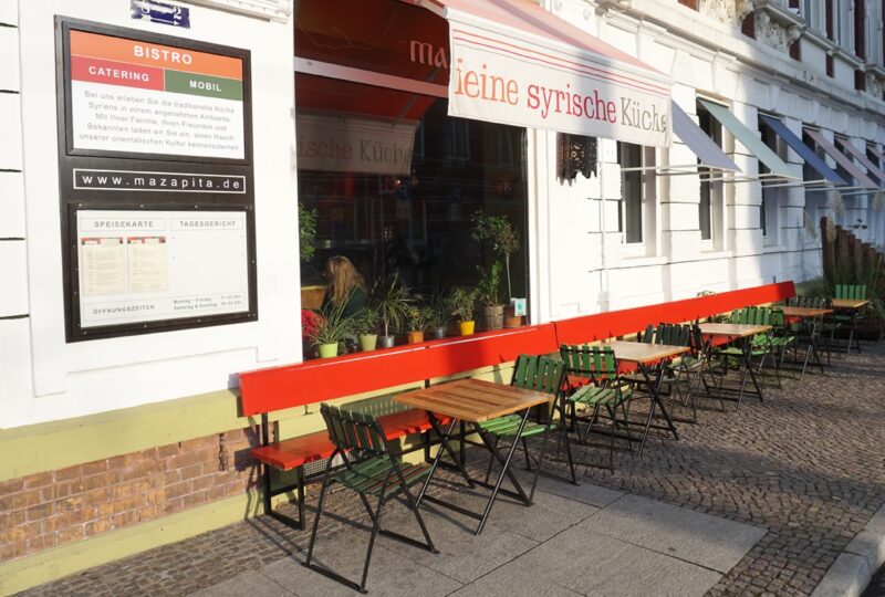 Empty outdoor seating area with green and wooden tables in front of a bistro, featuring a sign for catering services.