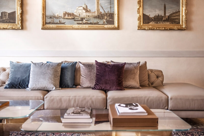 Elegant sofa with colorful pillows in a stylish room, featuring vintage paintings and a glass coffee table.