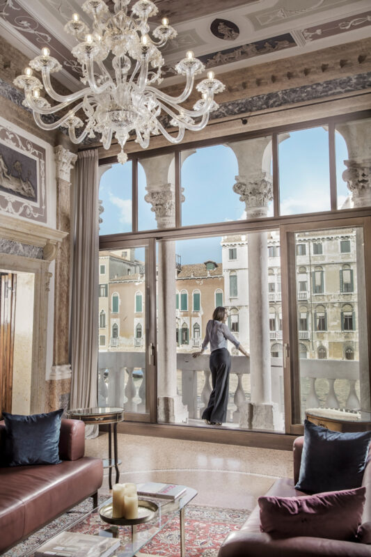Elegant interior of Palazzo Garzoni with a woman gazing out at Venetian architecture through large windows.