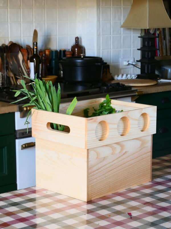 Wooden box with cut-out handles, filled with green vegetables, placed on a checkered table in a kitchen setting.