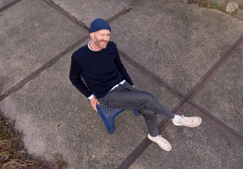 Man sitting casually on a blue mesh chair, wearing a navy sweater, gray pants, and a beanie, smiling at the camera.