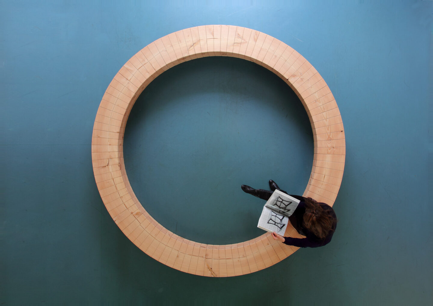 Circular wooden structure on a blue floor, with a person reading a book while standing inside the ring.
