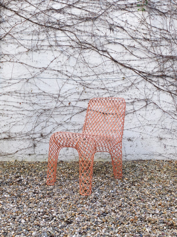 Orange mesh chair against a white wall with creeping vines, set on a gravel surface.