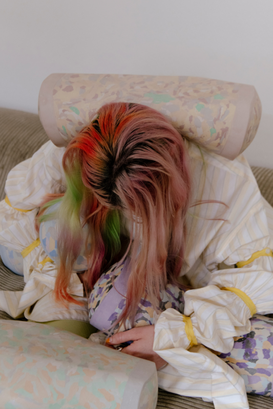 Person with colorful hair sitting on a couch, resting their head on a patterned neck roll, surrounded by soft cushions.