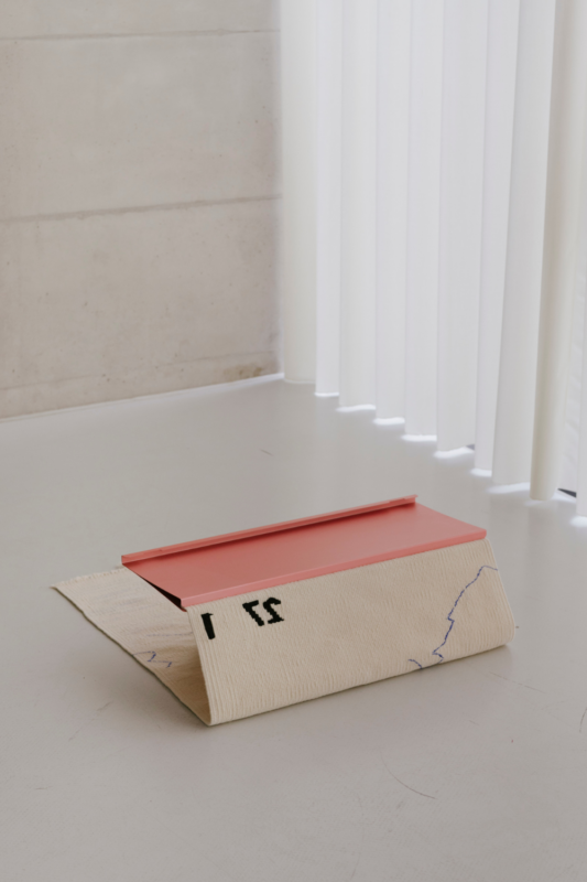 A folded tapestry with a red top and beige base, featuring abstract lines, placed on a light floor near vertical blinds.