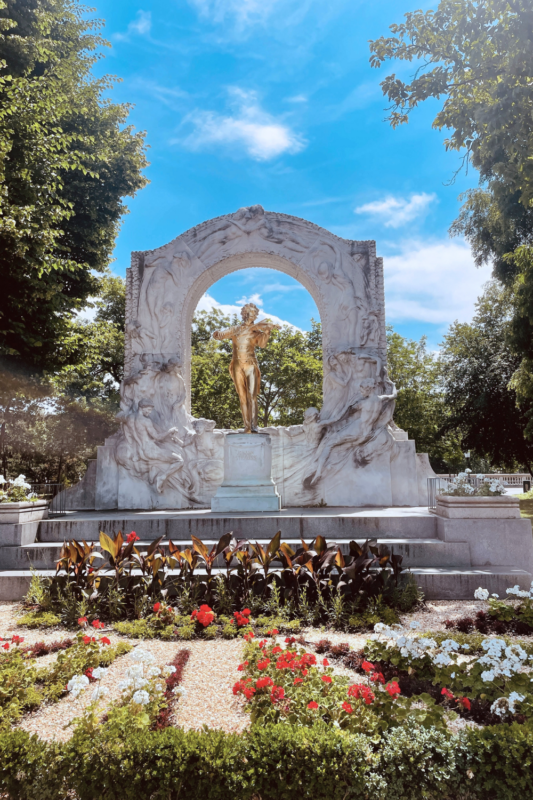 Golden statue surrounded by ornate stone arch, set in a garden with colorful flowers under a blue sky.