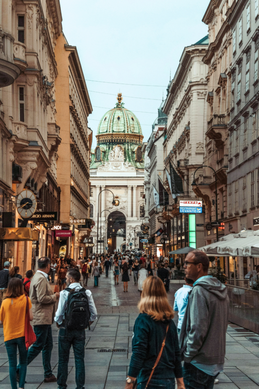 Busy street scene in Vienna, featuring a grand building with a green dome and people walking among shops and cafes.