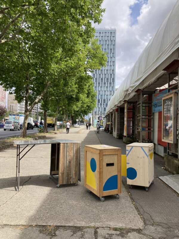 Wooden and colorful boxes on a sidewalk, with a modern building and people walking in the background.