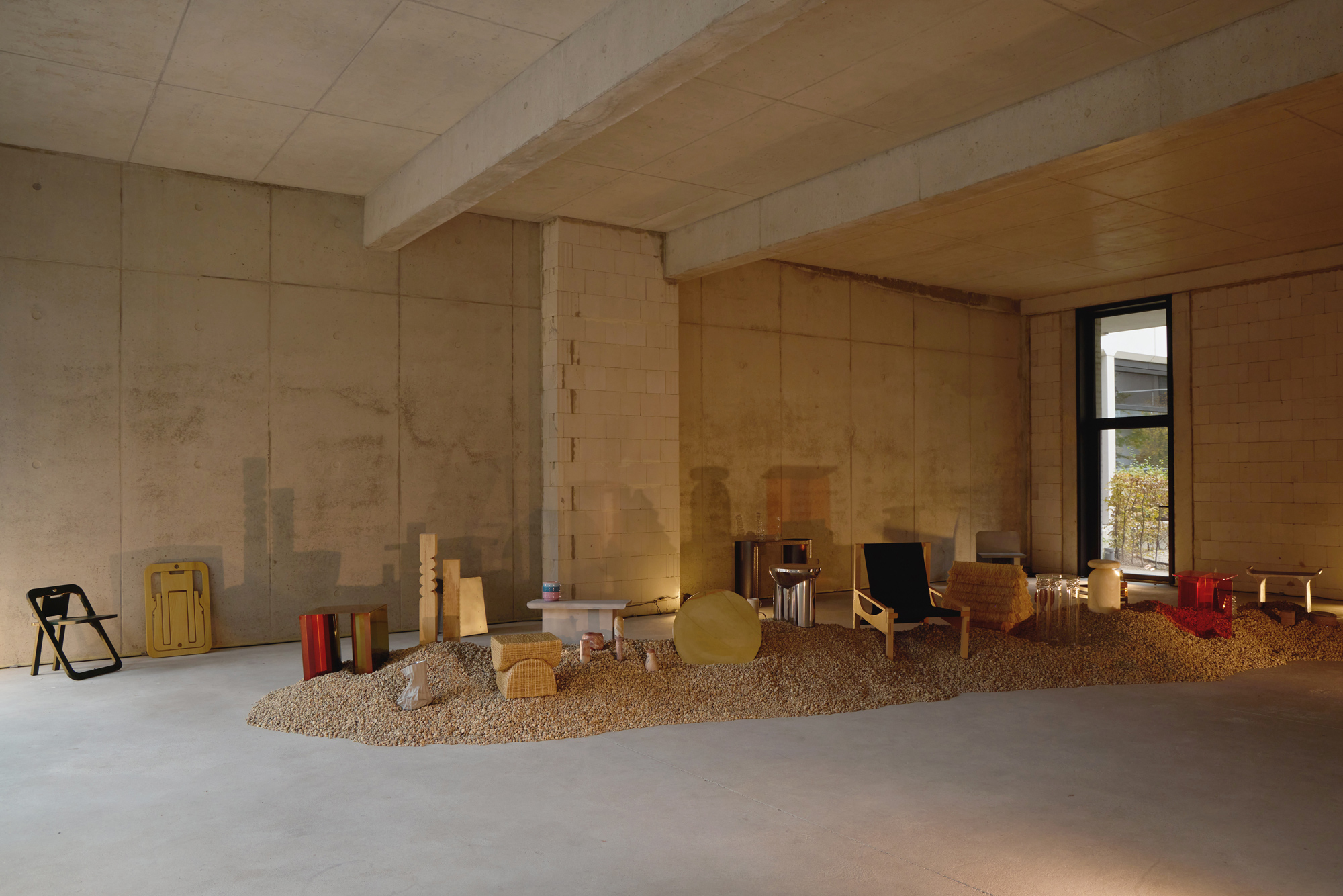 Exhibition space featuring various furniture pieces arranged on a gravel mound, with concrete walls and natural light.
