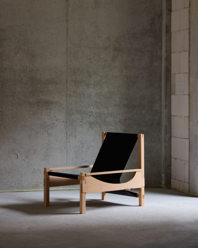 Minimalist wooden chair with a black fabric seat, positioned in a dimly lit room with a concrete wall.