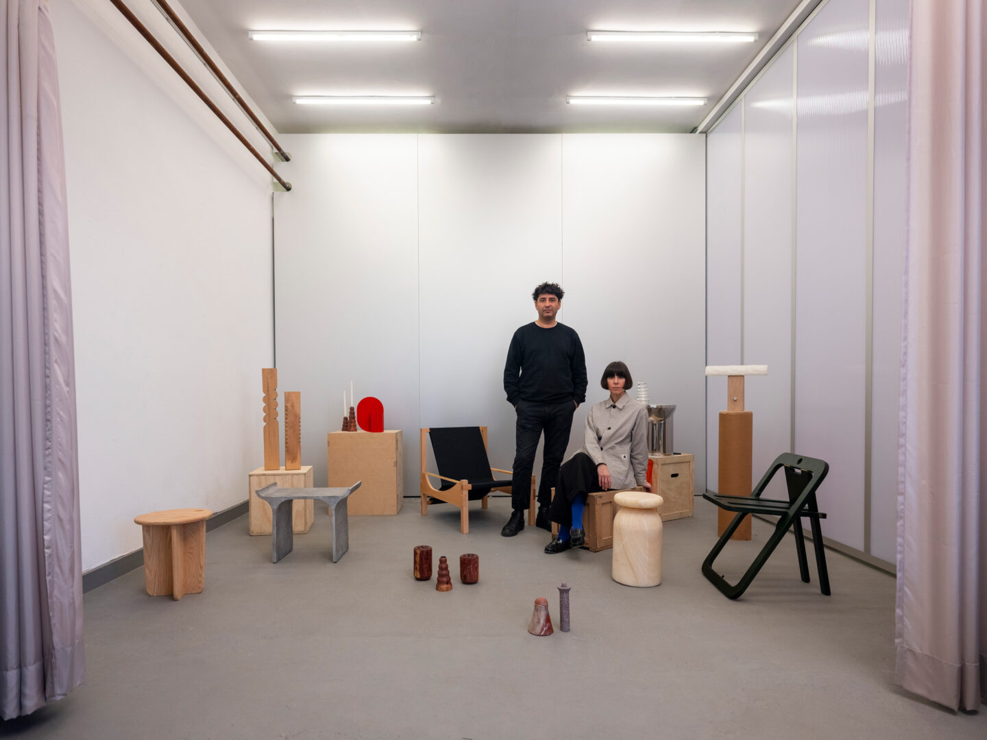 Two individuals stand among various artistic wooden furniture pieces in a minimalist gallery space.