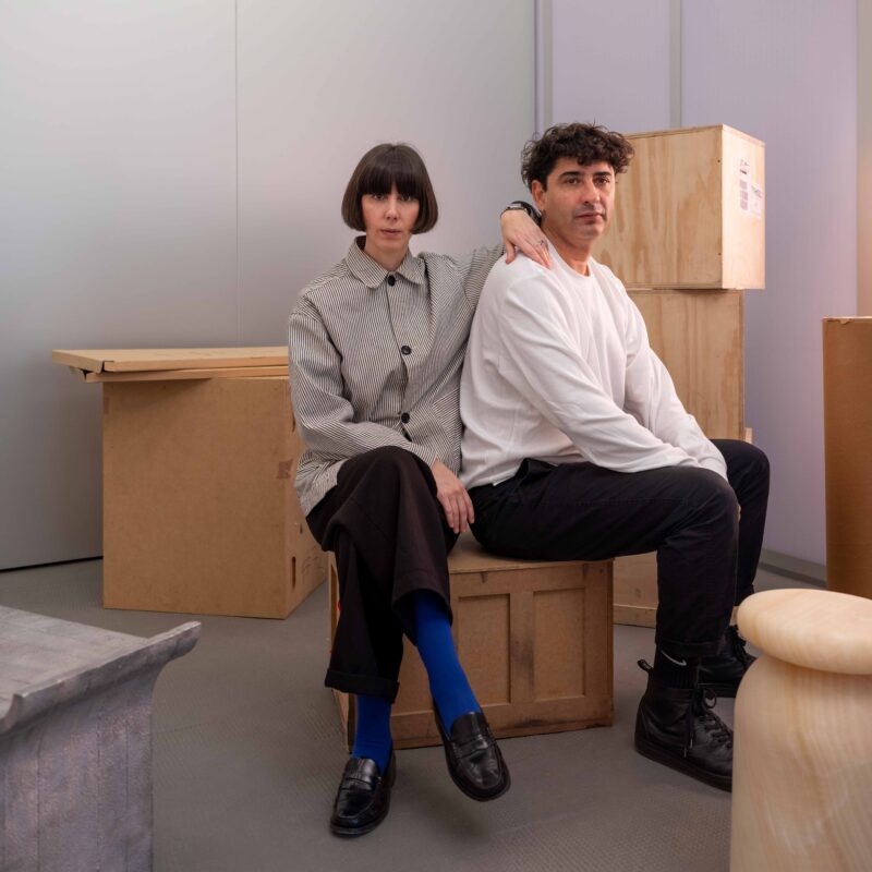 A woman in a striped shirt and a man in a white sweater sit on wooden boxes in a minimalist studio space.