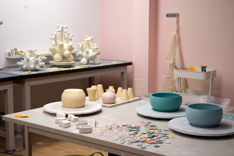 Ceramic studio with pastel-colored walls, featuring pottery pieces, tools, and colorful clay discs on tables.