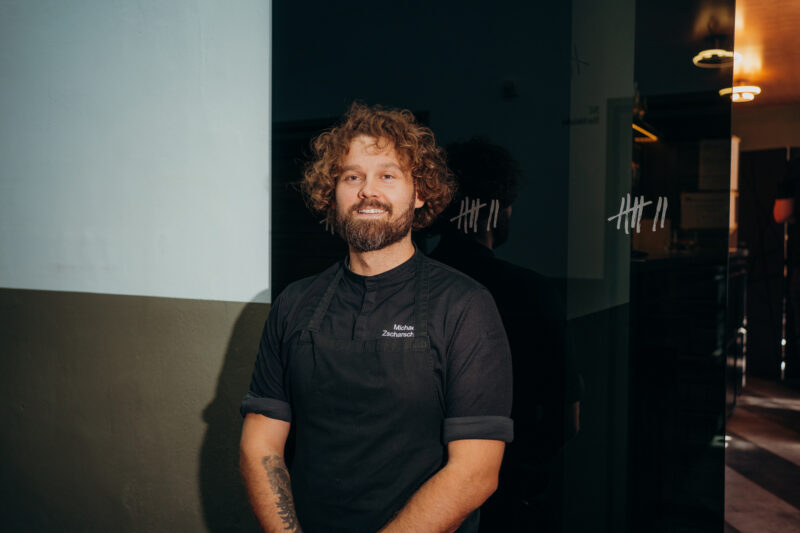 Smiling chef with curly hair stands in a dimly lit space, wearing a black apron, against a wall with markings.