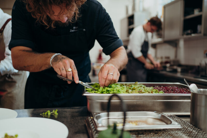 Chef preparing fresh greens in a kitchen, with another chef working in the background.