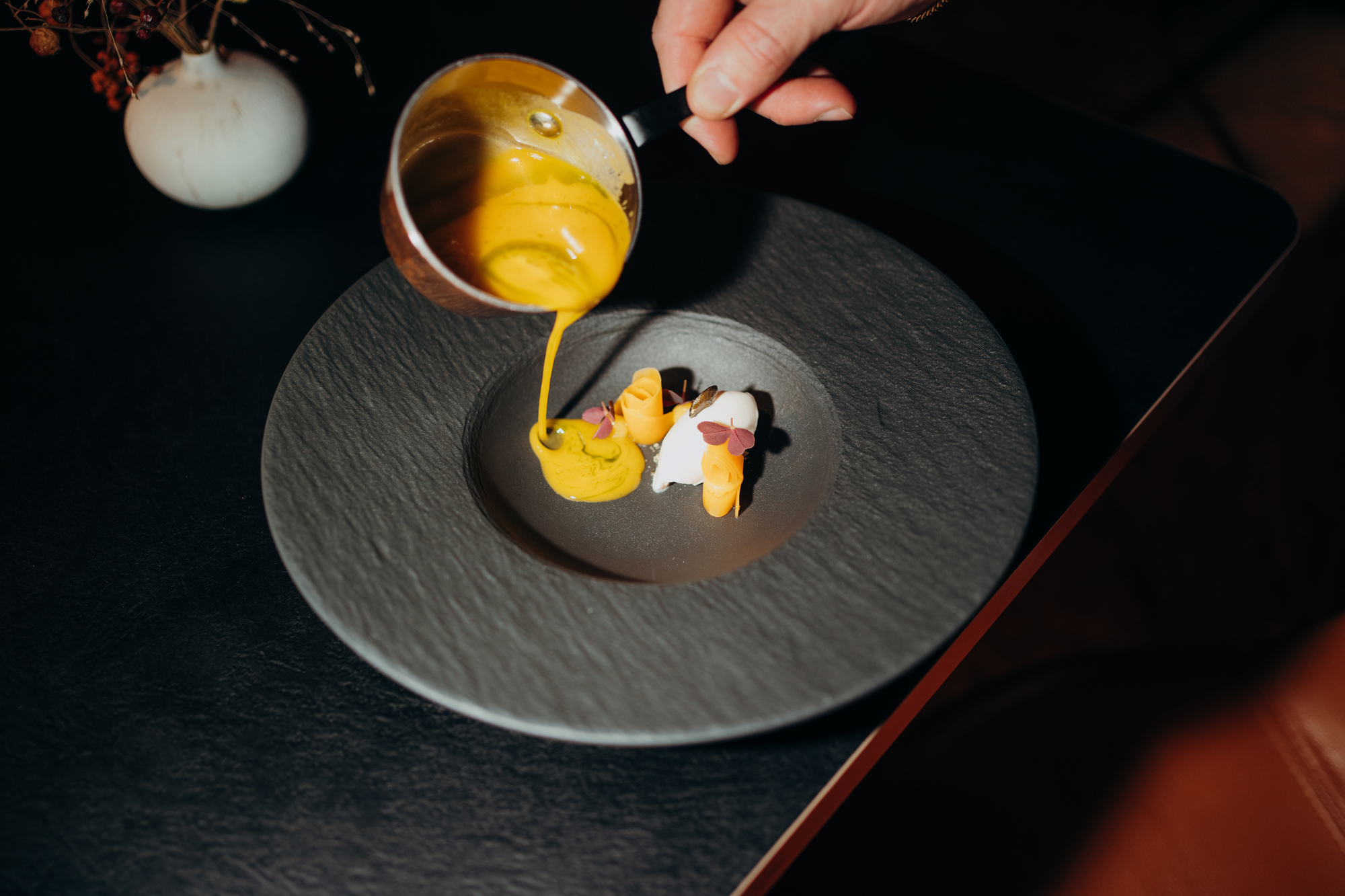 A hand pours yellow sauce over a plate with delicate food presentation, featuring small garnishes.