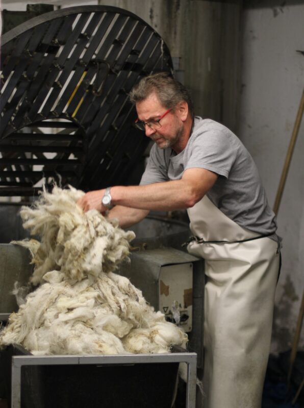 Man in gray shirt and apron handling a large pile of wool in a washing machine.