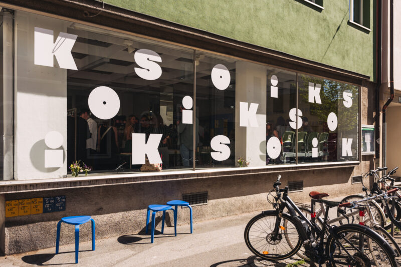 Storefront with large white letters spelling "KIOSK" on glass, blue chairs outside, and bicycles parked nearby.
