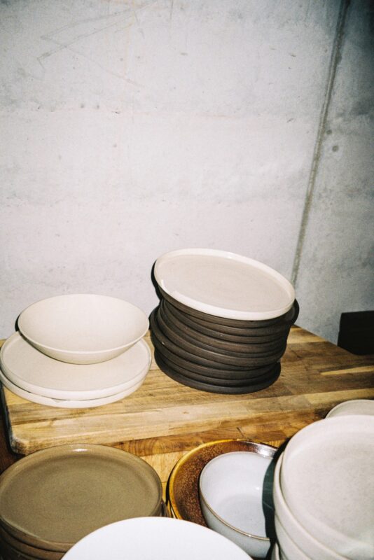 Stacked ceramic plates in various colors on a wooden surface, against a concrete wall.