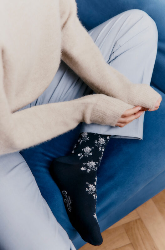 Model wearing navy floral socks, seated on a blue couch, with light-colored sweater and pale blue pants.