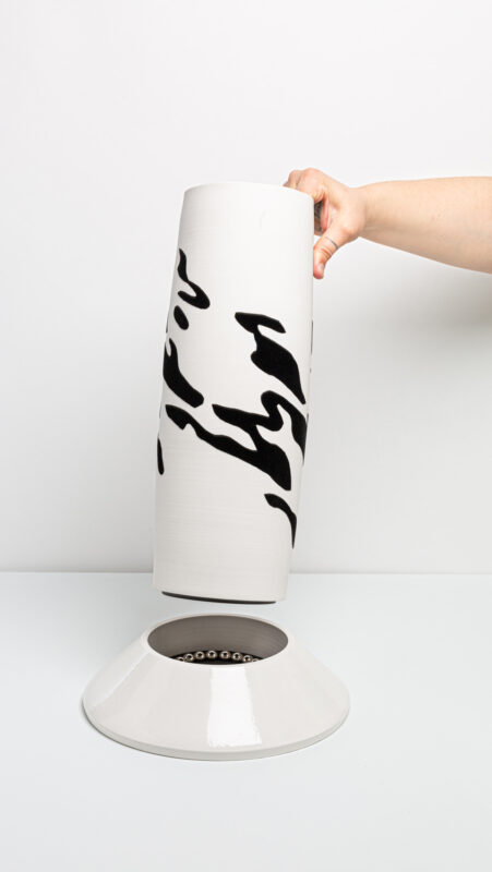 A hand holds a tall, white vase with black abstract patterns, positioned above a circular base.