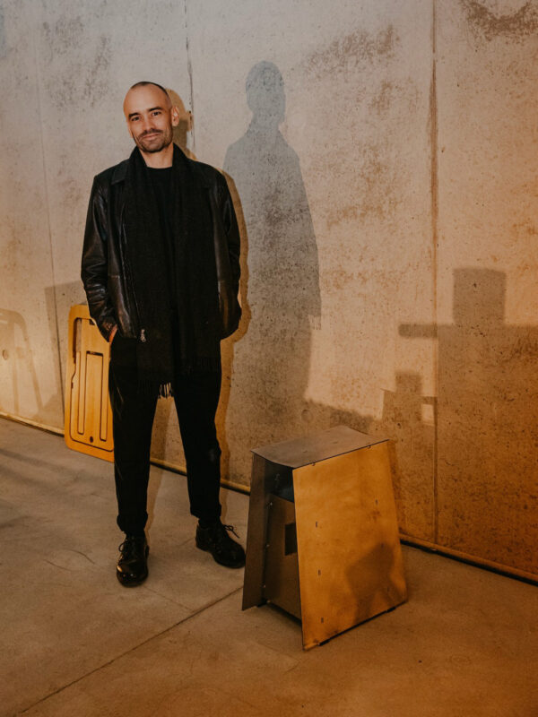 Man in a black leather jacket and scarf stands next to a metallic stool against a textured wall.