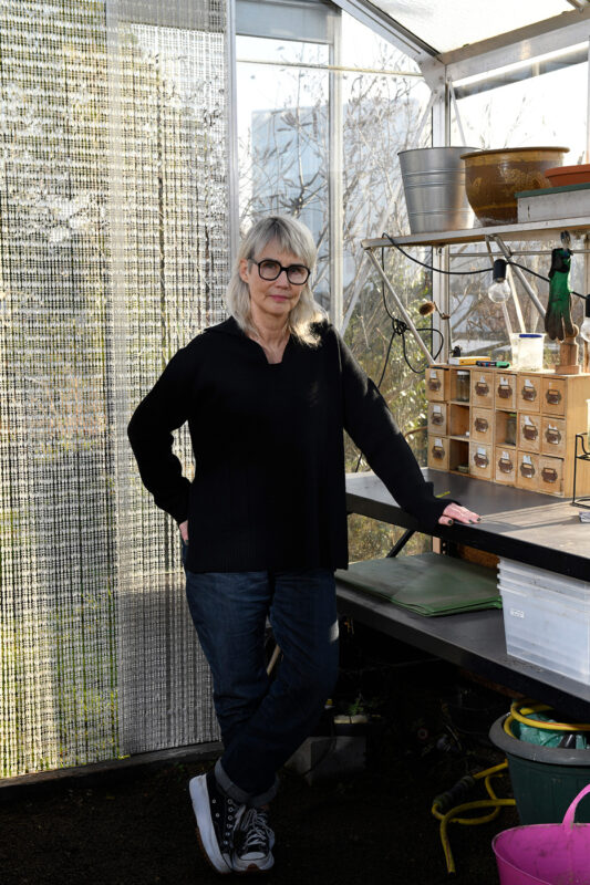 Woman with glasses in a greenhouse, leaning against a workbench with gardening tools and plants around her.
