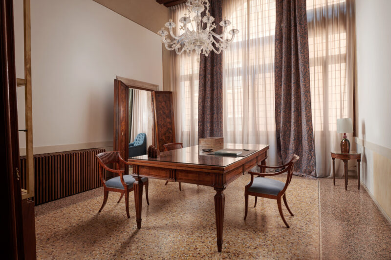 Elegant office space in Palazzo Garzoni featuring a wooden desk, two chairs, a chandelier, and large windows with drapes.