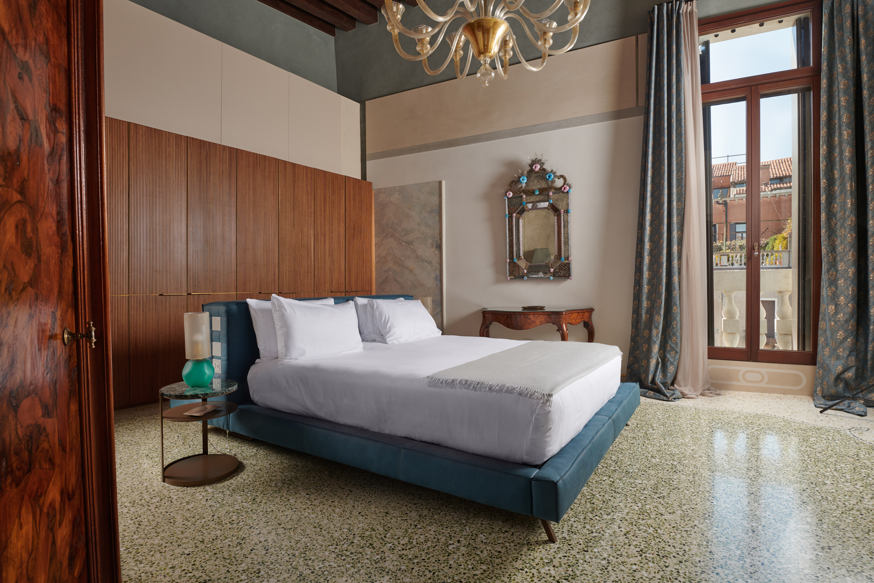 Modern bedroom in Palazzo Garzoni featuring a blue bed, elegant chandelier, and large windows with scenic views.
