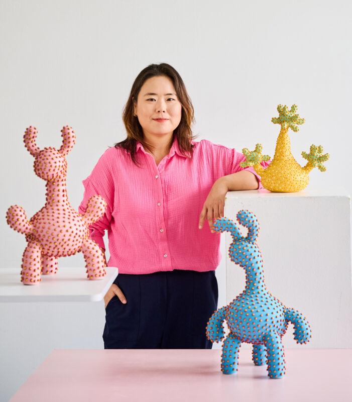 Portrait of Ahryun Lee in a pink shirt, standing beside colorful, textured sculptures on a white background.