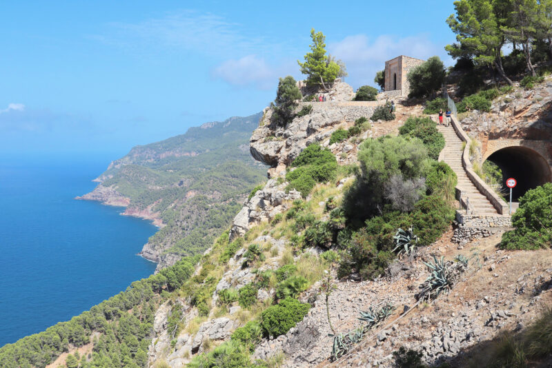 Scenic view of Serra de Tramuntana coastline with rocky cliffs, blue sea, and a historic building on the hillside.