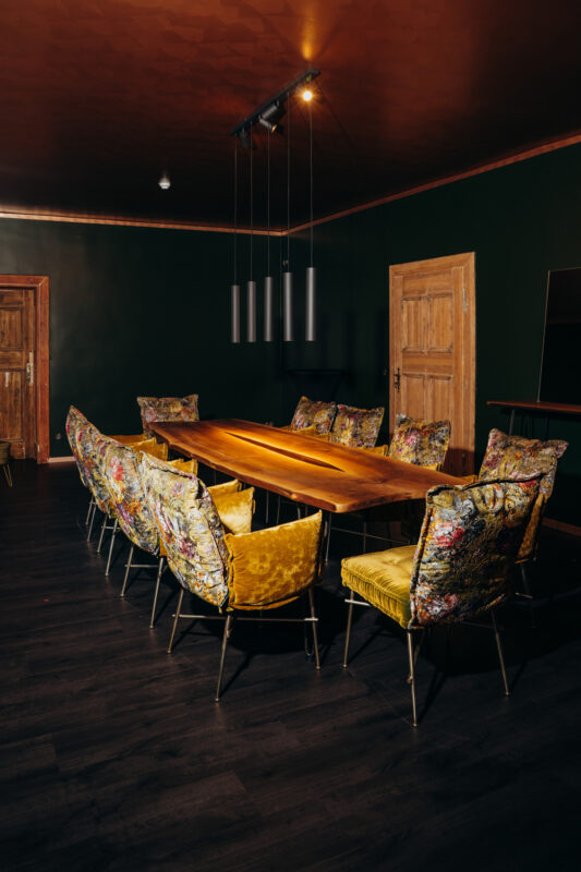 Private dining room with a long wooden table surrounded by colorful, patterned chairs against a dark green wall.