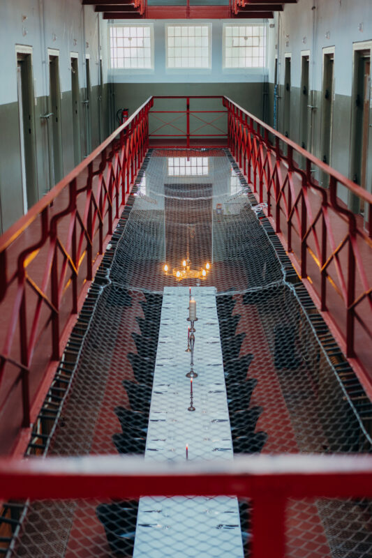 View of a prison cell block with a central walkway, red railings, and overhead lighting reflecting on the floor.