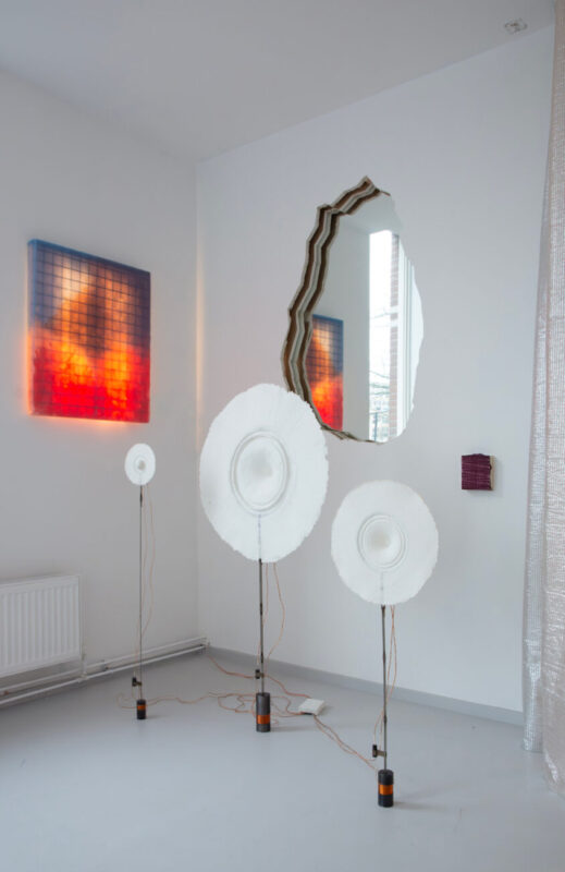 Three large, white circular sculptures on stands with wires, alongside a colorful wall artwork and a mirrored wall piece.