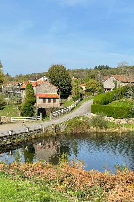 Rural landscape featuring houses with red roofs, a winding road, and a calm river under a blue sky.