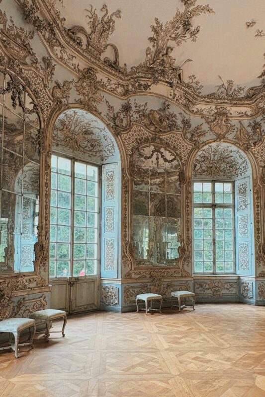 Ornate room with mirrored walls, large windows, and wooden flooring, featuring intricate decorations and two benches.