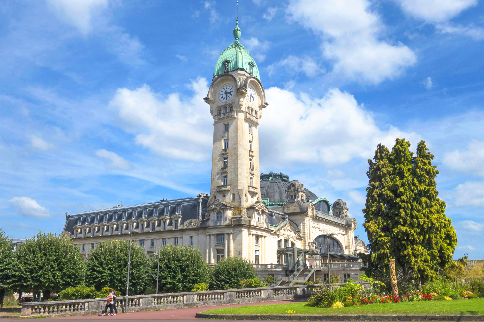 Historic building with a clock tower and green dome, surrounded by trees and a blue sky with clouds.