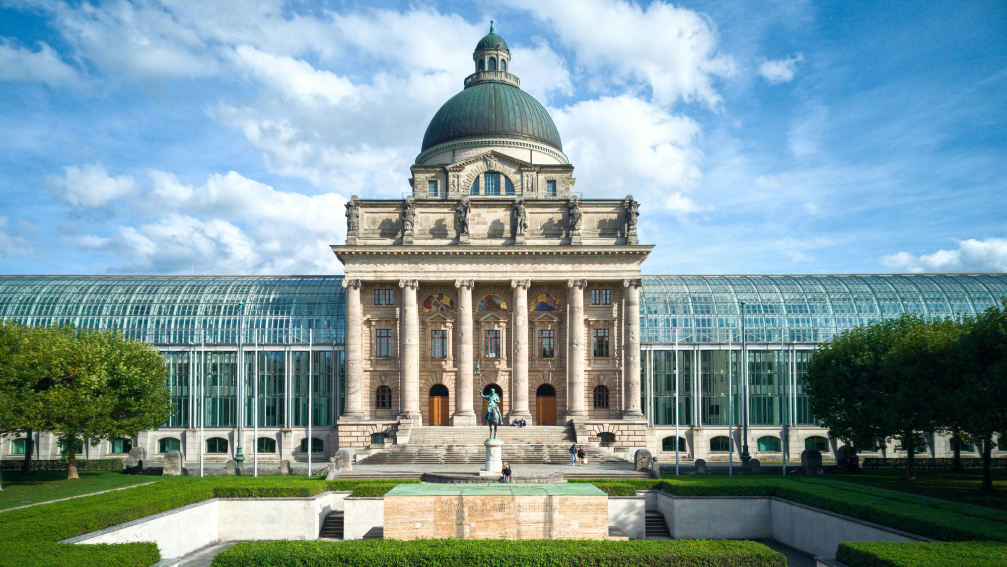 Historic building with a large dome, flanked by greenery and a statue in front, under a blue sky with clouds.