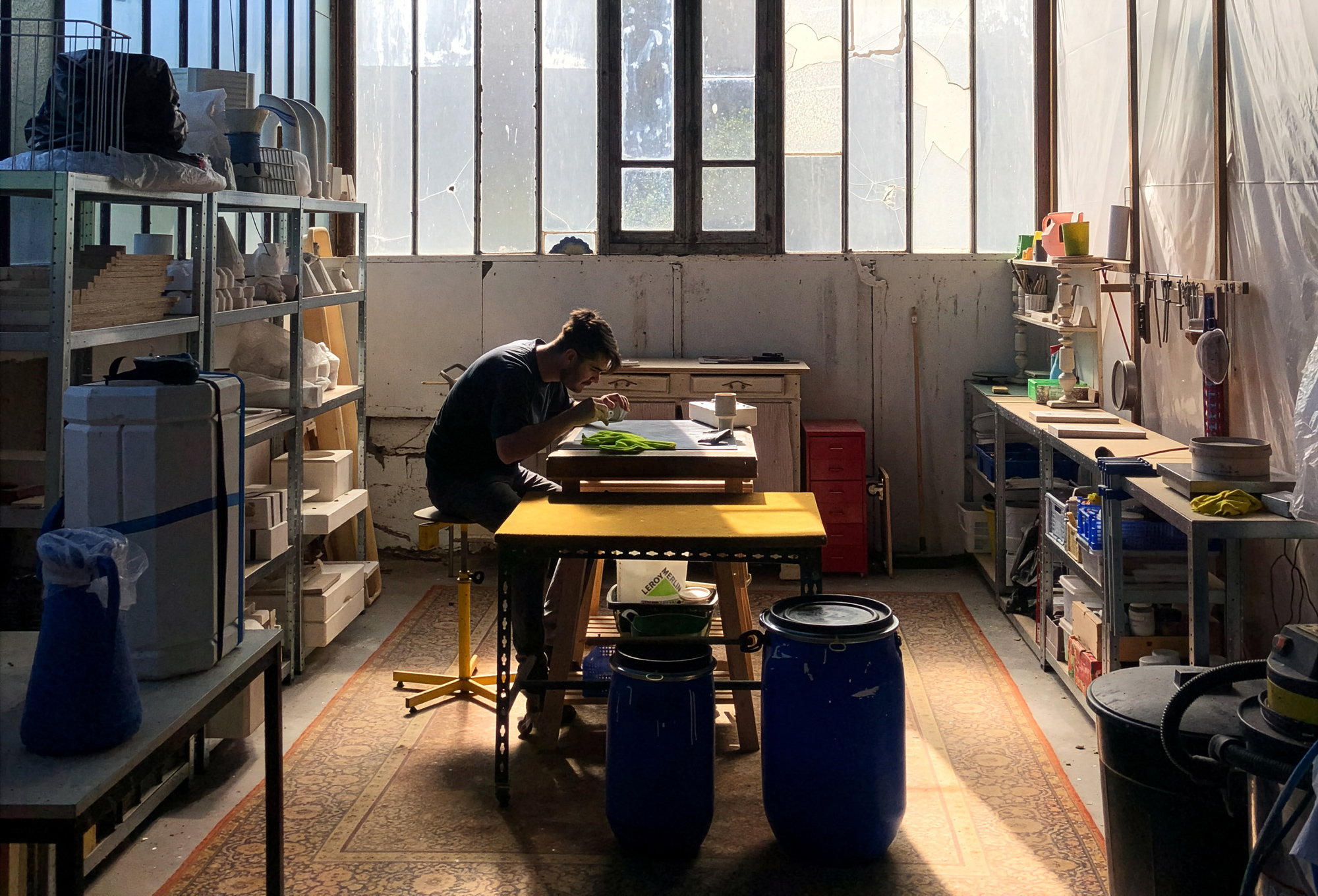 A man sits at a workbench in a sunlit workshop, focused on his task, surrounded by tools and materials.