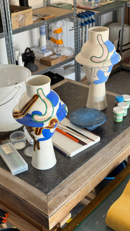 Two ceramic vases with colorful designs on a worktable, surrounded by art supplies in a workshop setting.