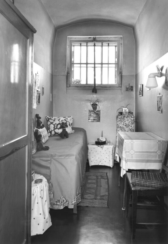 Prison cell interior featuring a bed, small table, and window, decorated with photos and personal items.