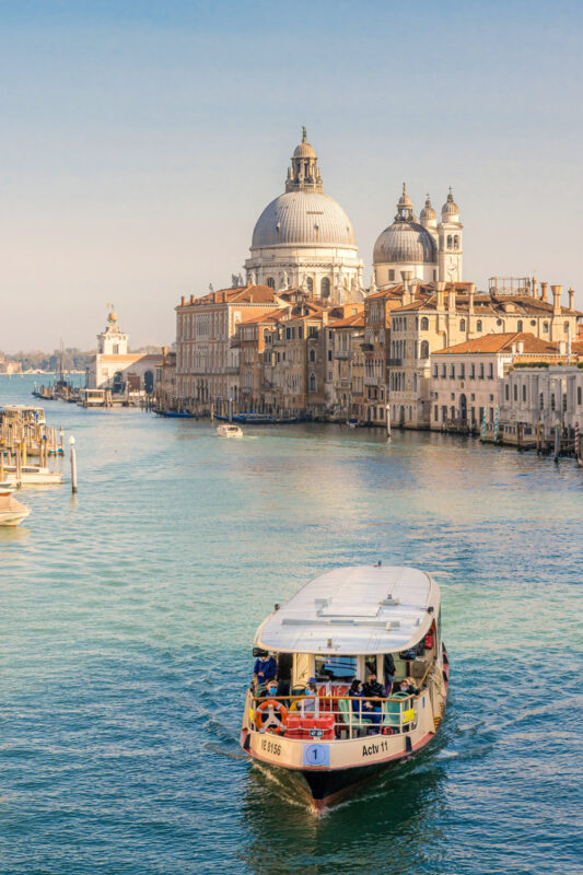 A boat navigates a serene canal in Venice, with historic buildings and domed churches lining the waterfront.