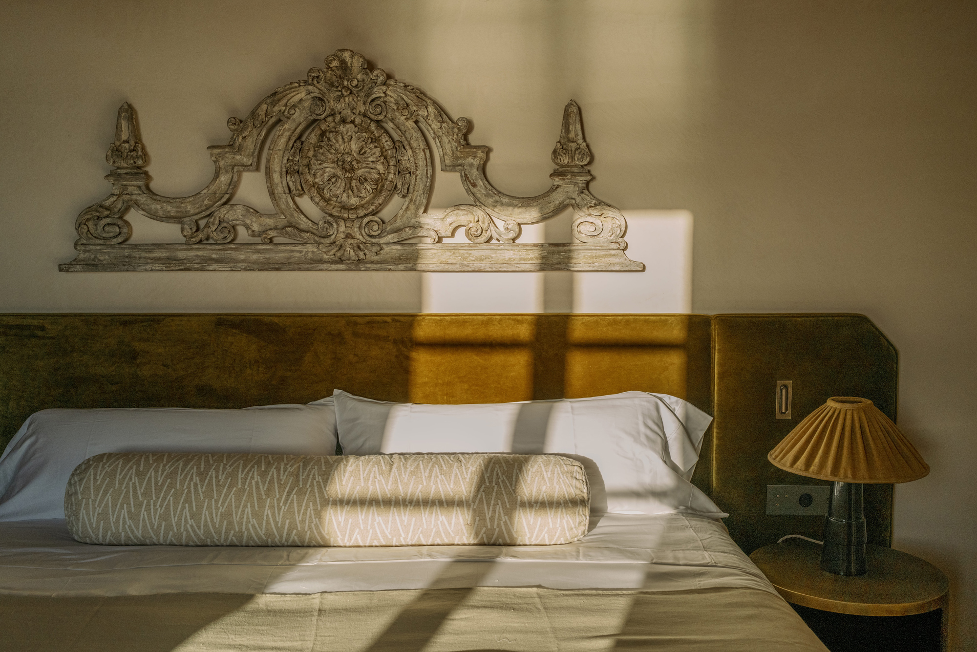 Elegant bedroom featuring a decorative headboard, a long pillow, and a stylish lamp, with soft shadows from sunlight.