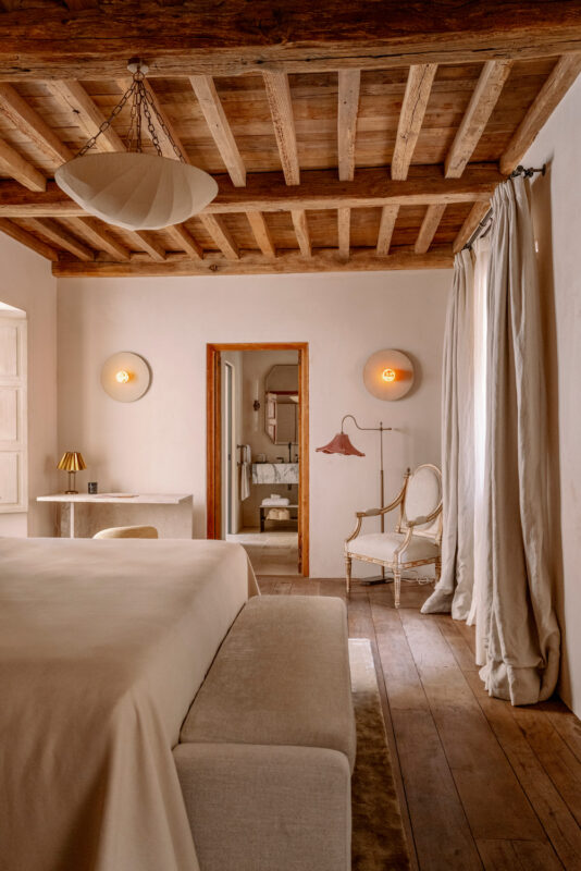 Elegant bedroom with wooden beams, light-colored walls, a plush bed, and a vintage chair near an open doorway to a bathroom.